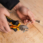 A person using a pair of Tolsen diagonal cutting pliers to cut a white electrical cable on a wooden surface