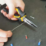 Close-up of hand using Tolsen 6-inch long nose pliers to cut thin metal wire on a dark surface